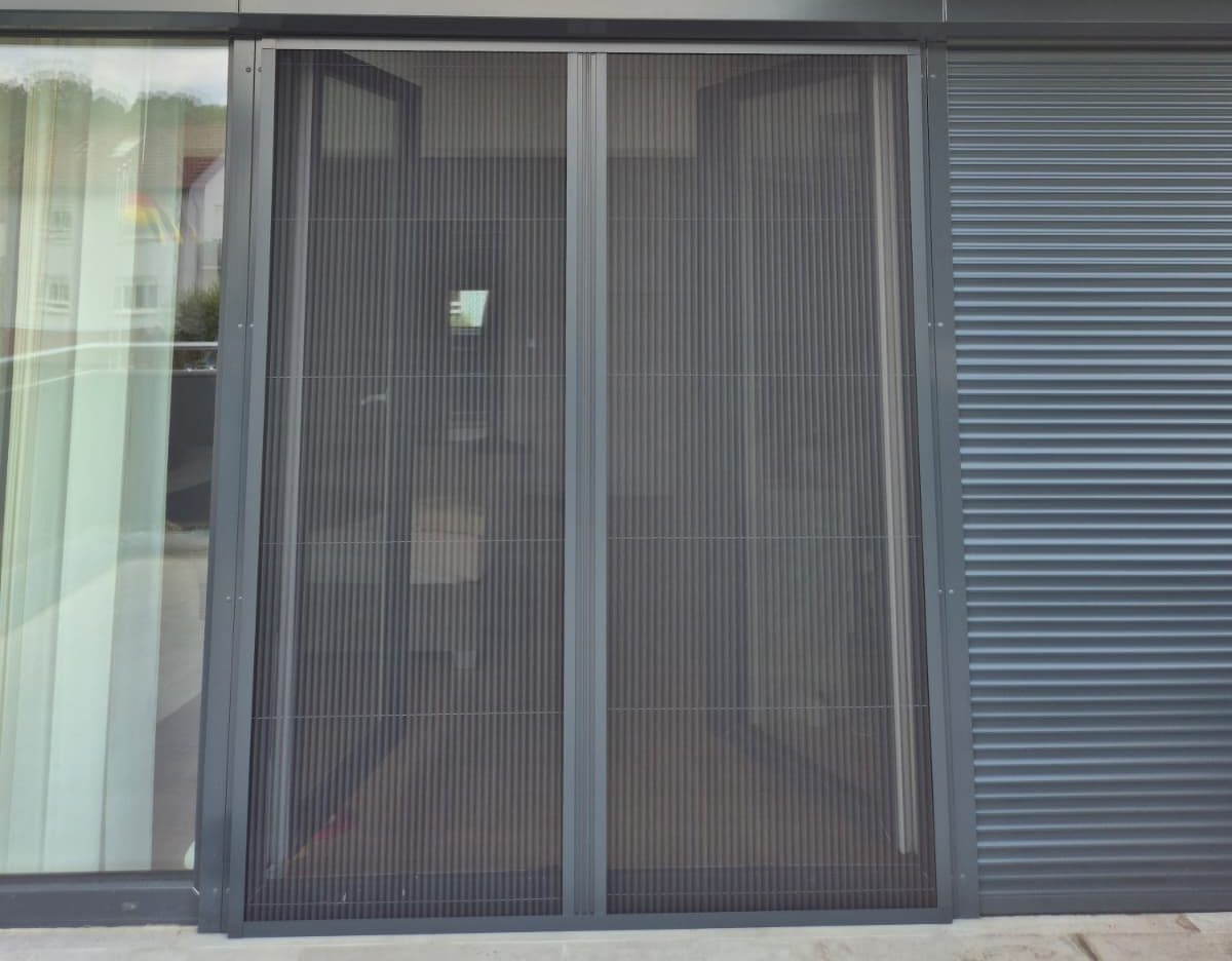 Fenster mit Gitter und grauer Außenwand, darunter ein grauer Rollladen.